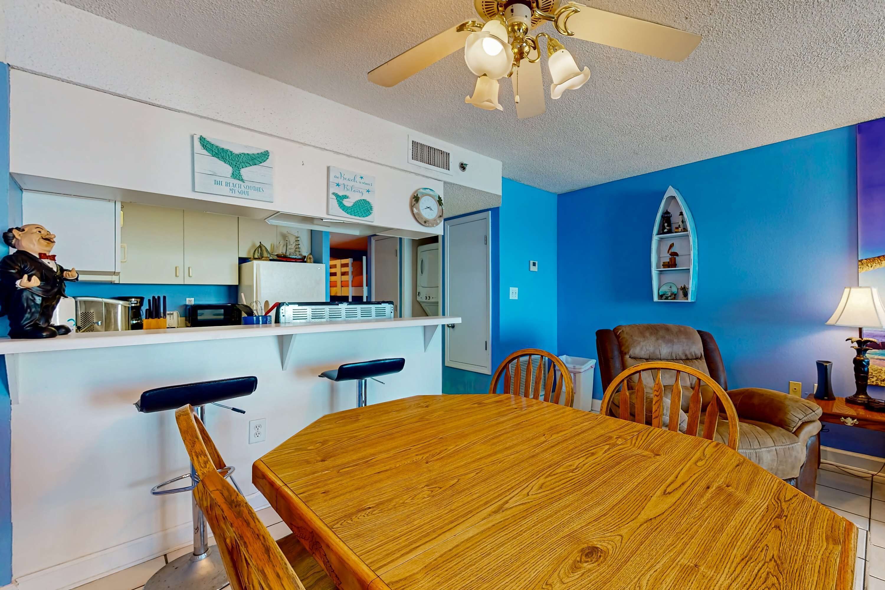 A room with a wooden dining table, bar stools, a ceiling fan, a microwave, a refrigerator, and a blue accent wall.