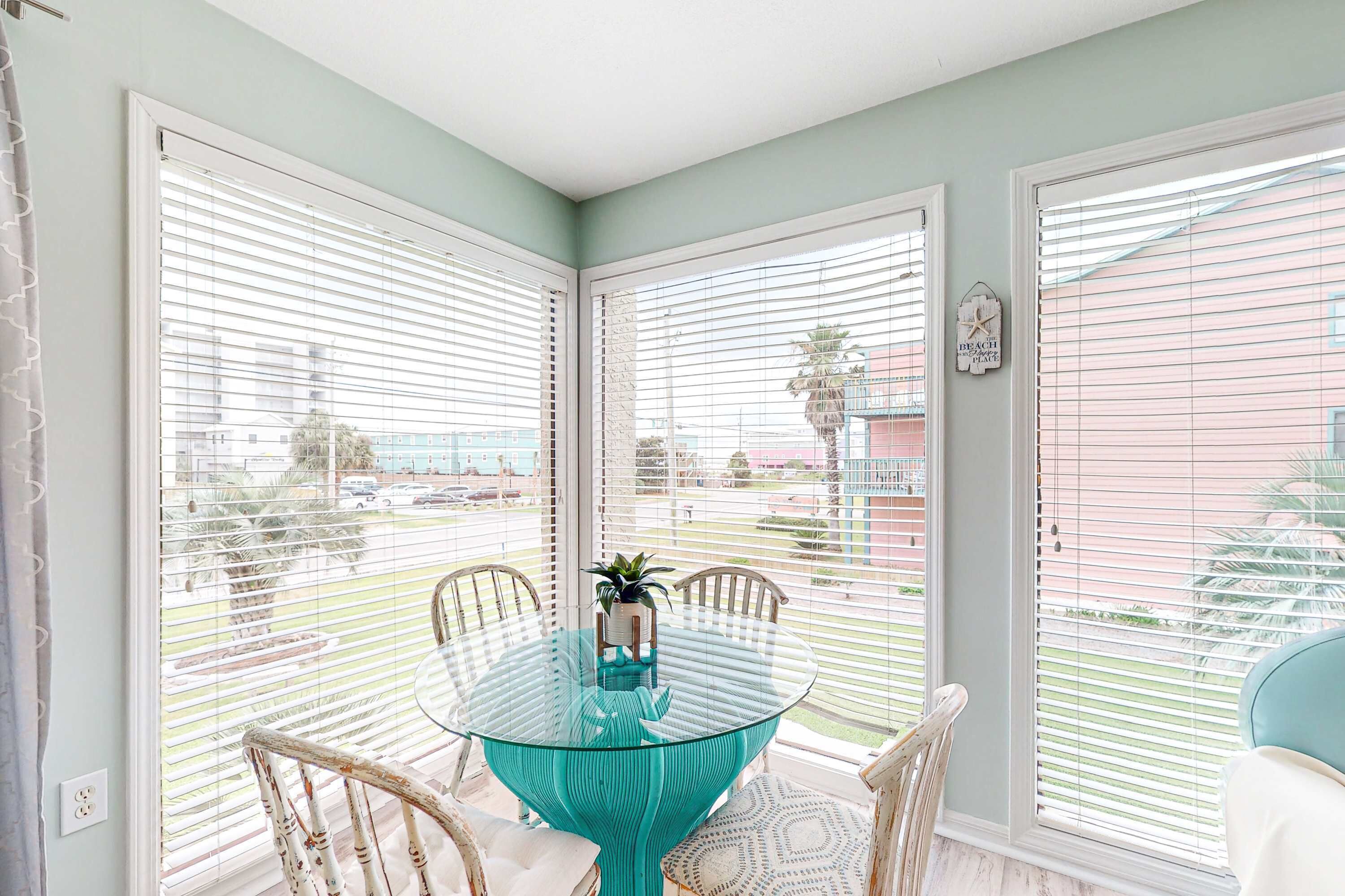 A dining area with a glass table surrounded by chairs, large windows with blinds, and a view of the outside.