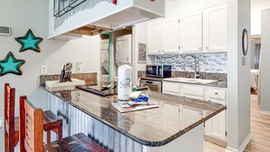 A kitchen with white cabinets, a granite countertop, a microwave, and a sink. - Centrally Located Oasis | Pool & Covered Balcony | Walk 1 Block to Beach (Gulf Shores)