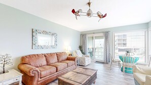 A living room with a brown leather sofa, a wooden coffee table, a dining table with chairs, and a balcony with a view. - Centrally Located Oasis | Pool & Covered Balcony | Walk 1 Block to Beach (Gulf Shores)