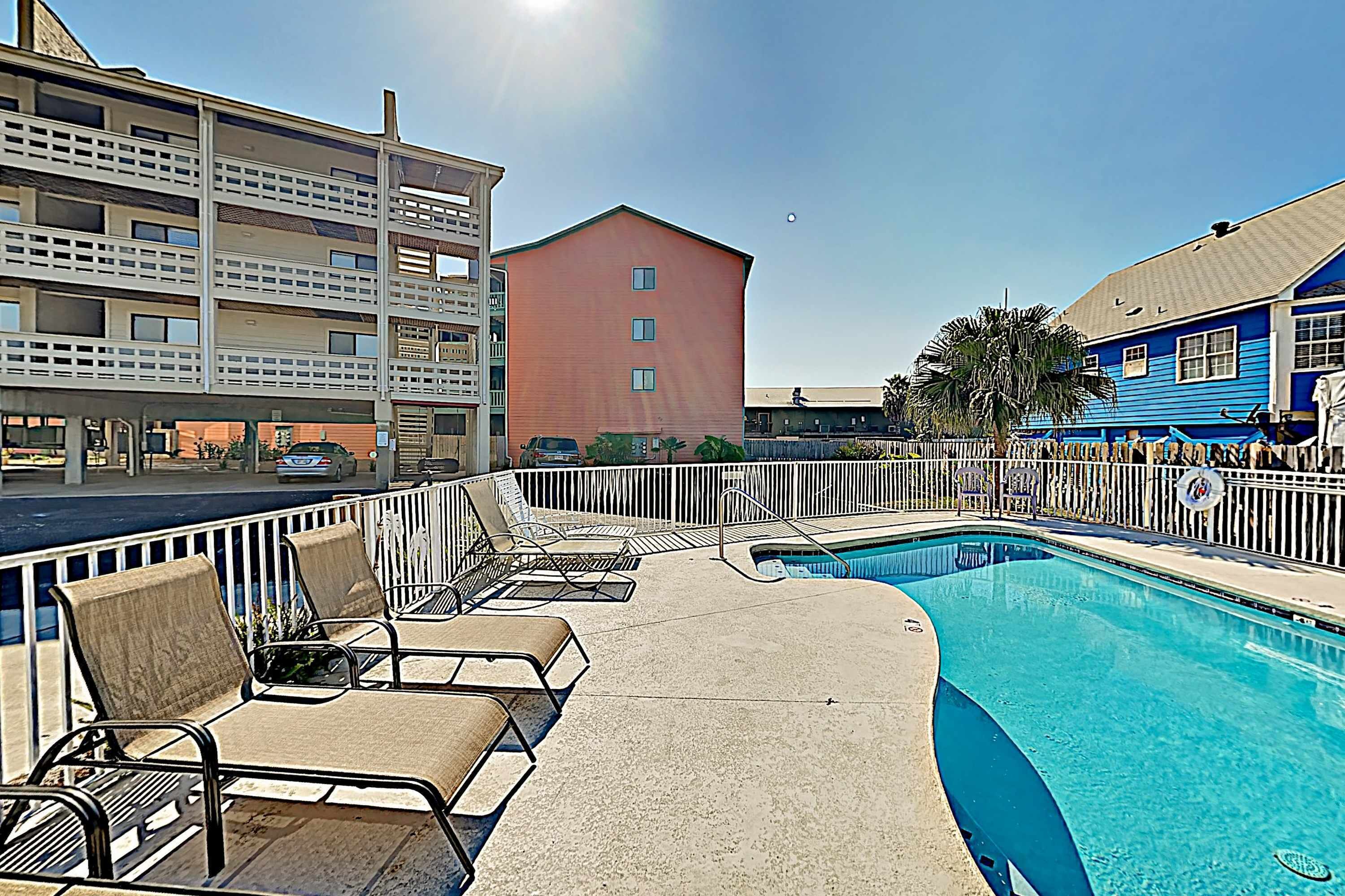 A pool area with lounge chairs, a clear pool, and a building with balconies in the background.