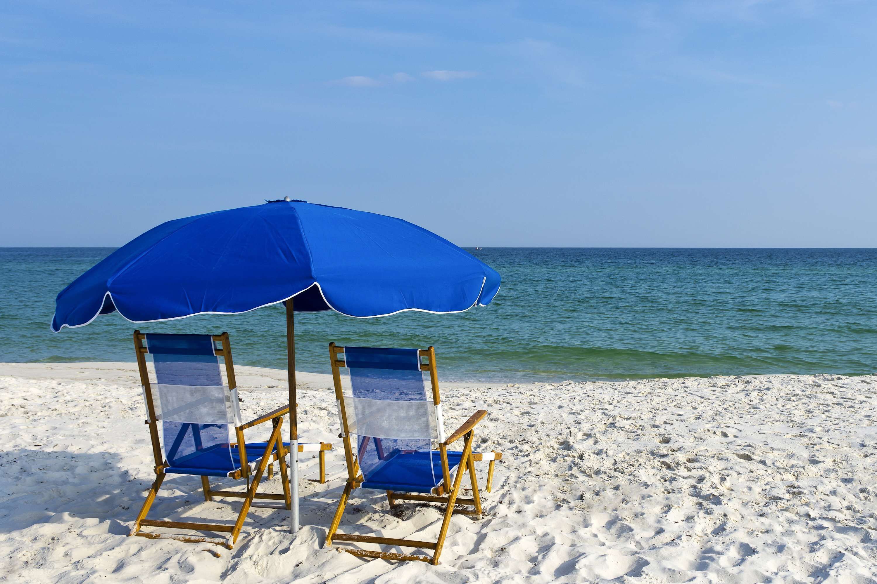 To strandstole under en blå parasol på en sandstrand med oceanen i baggrunden.