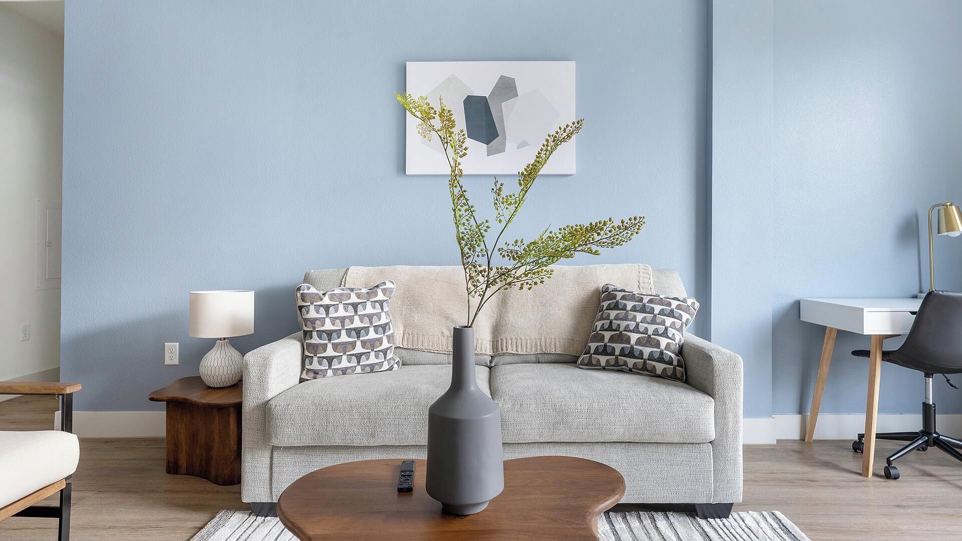 A living room with a sofa, a coffee table, a lamp, a vase with branches, and a desk with a chair.