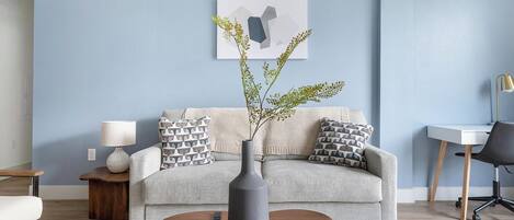 A living room with a sofa, a coffee table, a lamp, a vase with branches, and a desk with a chair.