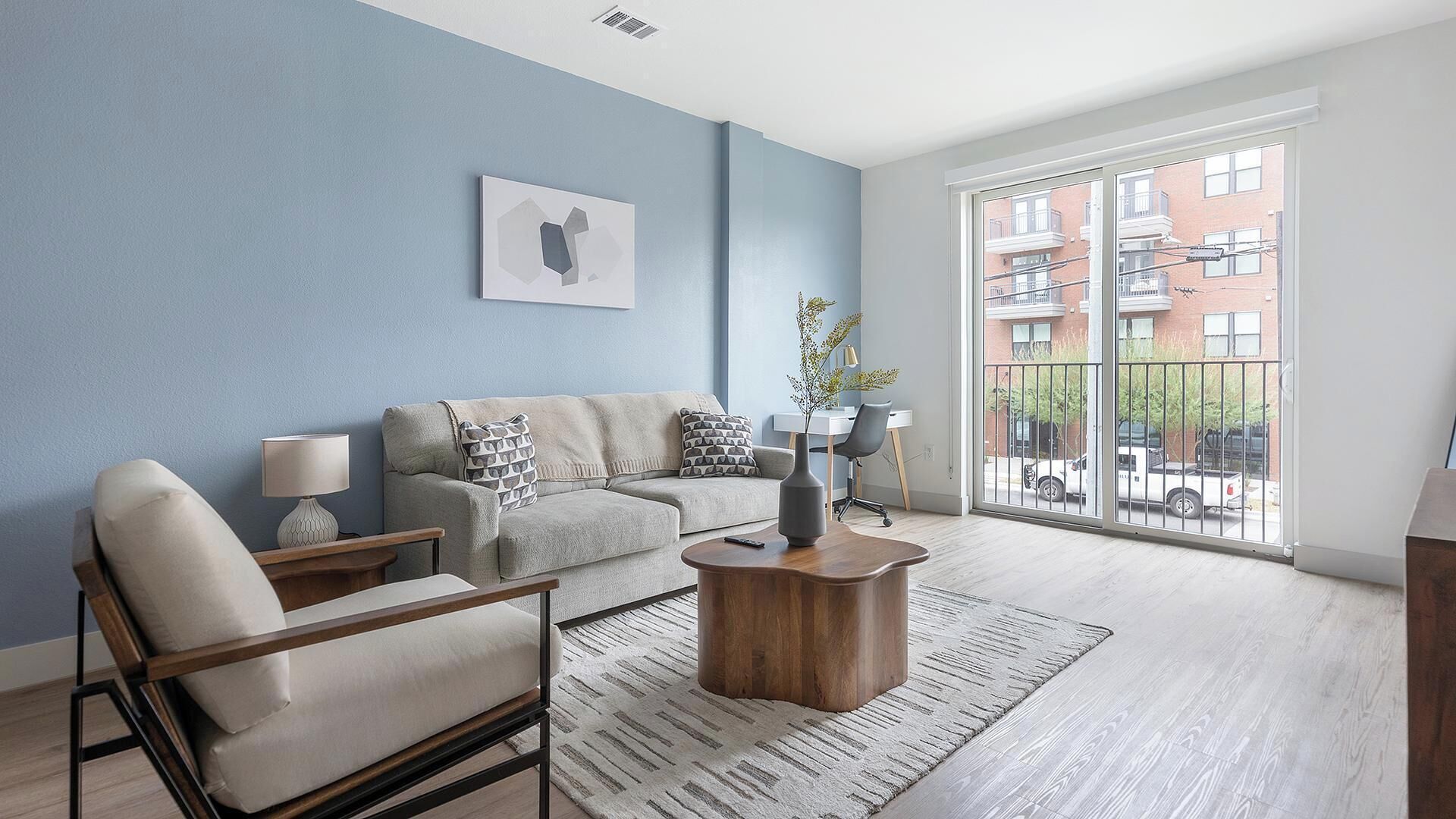 A modern living room with a sofa, armchair, coffee table, and a large window with a view of buildings.