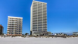 Een hoog hotel met verschillende balkons, een strand op de voorgrond en een helderblauwe lucht.