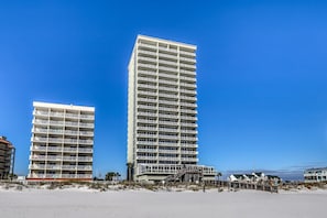 A tall hotel with multiple balconies, a beach in the foreground, and a clear blue sky. - Close to Beach The Colonnades, Stellar Gulf-Front Gem, Pool, Sauna, Gym (Gulf Shores)
