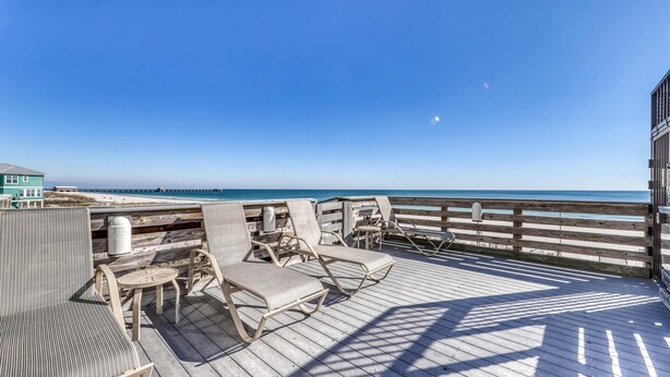 Een houten terras met lounge stoelen en tafels, met uitzicht op een strand en de oceaan.