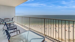Ein Balkon mit Glastisch und Stühlen, der auf einen Strand mit Sonnenschirmen und Personen mit Blick blickt.