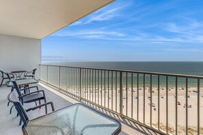 Ein Balkon mit Glastisch und Stühlen, der auf einen Strand mit Sonnenschirmen und Personen mit Blick blickt.