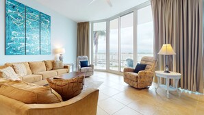 A spacious living room with a large window, a ceiling fan, and a view of the outdoors. - Brilliant water view condo with multiple pools, lazy river & internet (Orange Beach)
