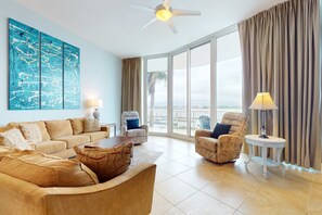A spacious living room with a large window, a ceiling fan, and a view of the outdoors. - Brilliant water view condo with multiple pools, lazy river & internet (Orange Beach)
