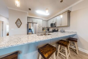 A modern kitchen with granite countertops, stainless steel appliances, and wooden bar stools. - Dog-Friendly Condo with Shared Pool, Central AC, & Free High-Speed WiFi (Gulf Shores)