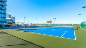 A tennis court with a net, surrounded by a fence, and a building with balconies in the background. - Inviting Condo w/ Shared Pools, Hot Tub, Tennis, & Beach Access (Gulf Shores)