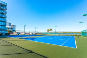 A tennis court with a net, surrounded by a fence, and a building with balconies in the background. - Inviting Condo w/ Shared Pools, Hot Tub, Tennis, & Beach Access (Gulf Shores)