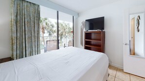 A hotel room with a large bed, a television on a wooden stand, a balcony with a view of palm trees, and a door with a mirror. - Inviting Condo w/ Shared Pools, Hot Tub, Tennis, & Beach Access (Gulf Shores)