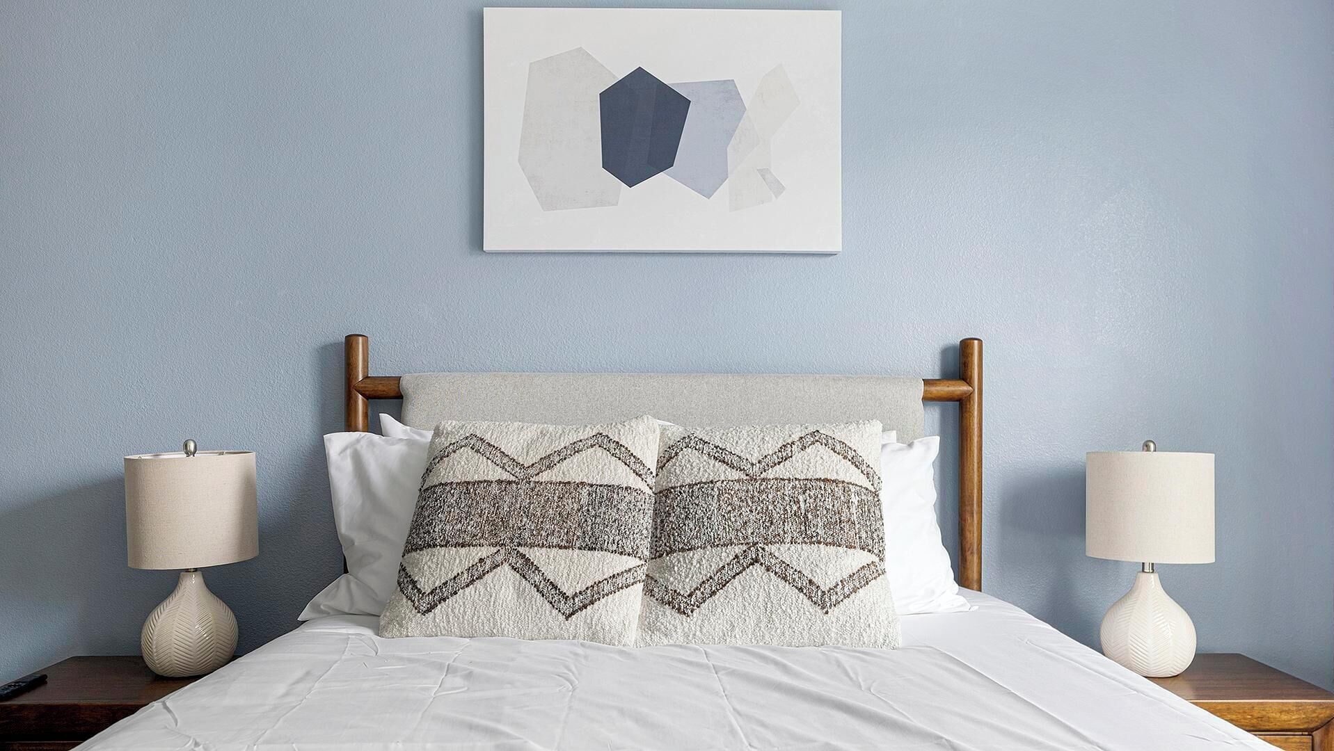 En seng med hvide sengetæger, to natborde med lamper, et træhovedgærde og et geometrisk abstrakt billede på væggen.
