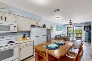 A kitchen with white cabinets, a microwave, and a refrigerator. There is a dining table with blue placemats and wine glasses. - Partial lagoon-view charming condo with beach access minutes away (Gulf Shores)