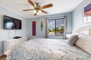 Une chambre à coucher comprenant un lit, un ventilateur de plafond, une télévision, une commode et une fenêtre avec des stores.