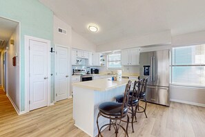 A modern kitchen with a central island, stainless steel appliances, and a dining area with four chairs. - Spacious, Gulf-front vacation home with covered deck & beach access (Gulf Shores)