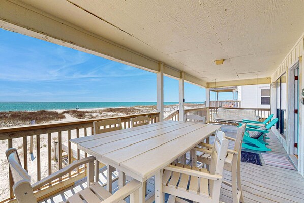 A covered outdoor area with a wooden table and chairs, a view of the beach, and a building with a balcony. - Spacious, Gulf-front vacation home with covered deck & beach access (Gulf Shores)