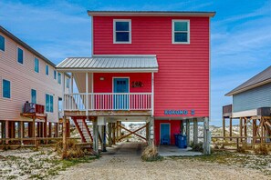 A red building with a balcony and a blue door, surrounded by wooden structures. - Memorable & group-friendly house w/Gulf views and private sun deck (Gulf Shores)