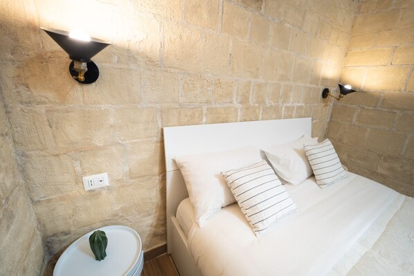 A bed with white linens and striped pillows, a round bedside table with a green object, and two wall-mounted light fixtures against a stone wall.