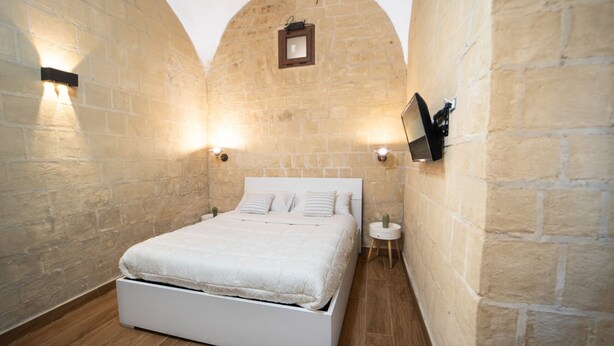 Une chambre avec un lit blanc, des murs en pierre et deux lampes fixées au mur.