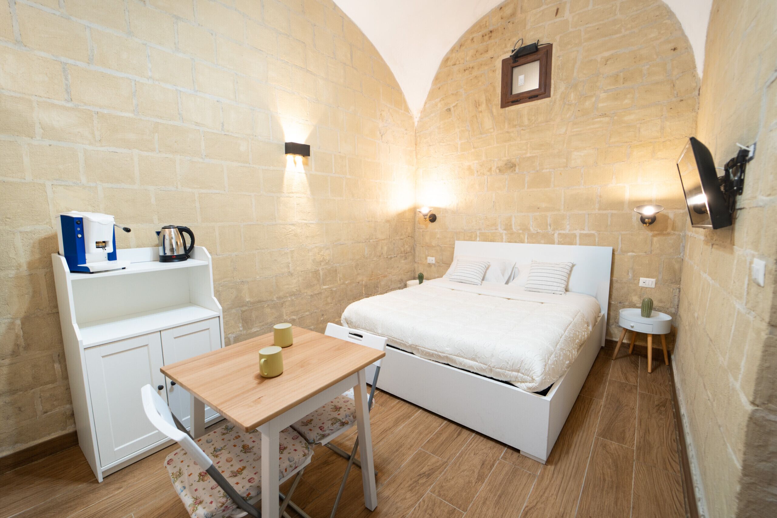 A bedroom with a bed, a desk, and a chair. There are two wall-mounted lights and a small round table with a plant.