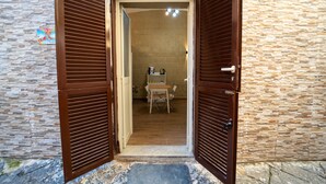 Une porte surmontée de volets en bois qui donne sur une pièce dont le sol est recouvert de carreaux, et qui contient une table et une chaise.