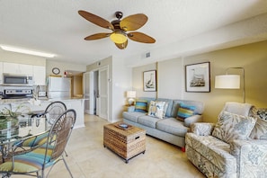 A living room with a sofa, chairs, a coffee table, a ceiling fan, and a kitchen area with appliances. - Gulf front condo with community outdoor and indoor pools, hot tub, and sauna (Gulf Shores)