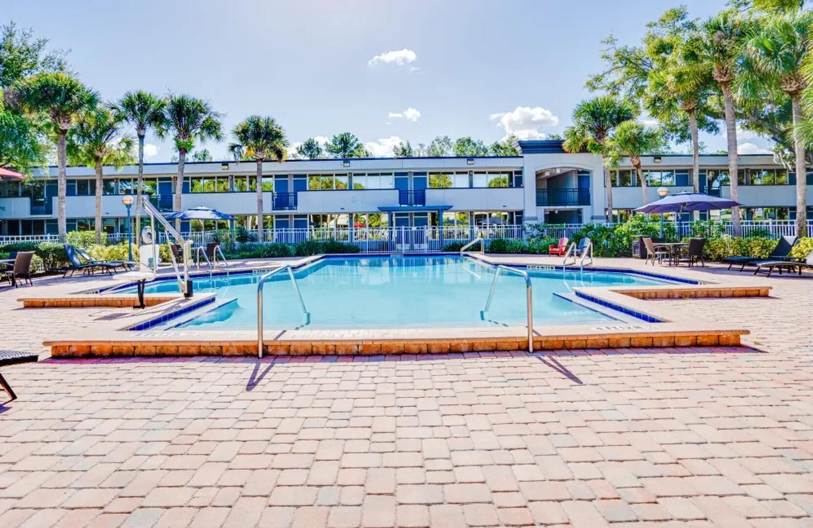 A swimming pool with a tiled deck, surrounded by palm trees and a building with multiple windows.