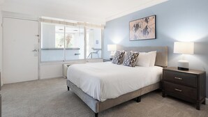 A bedroom with a bed, bedside tables, a nightstand, a window with blinds, and a painting on the wall.