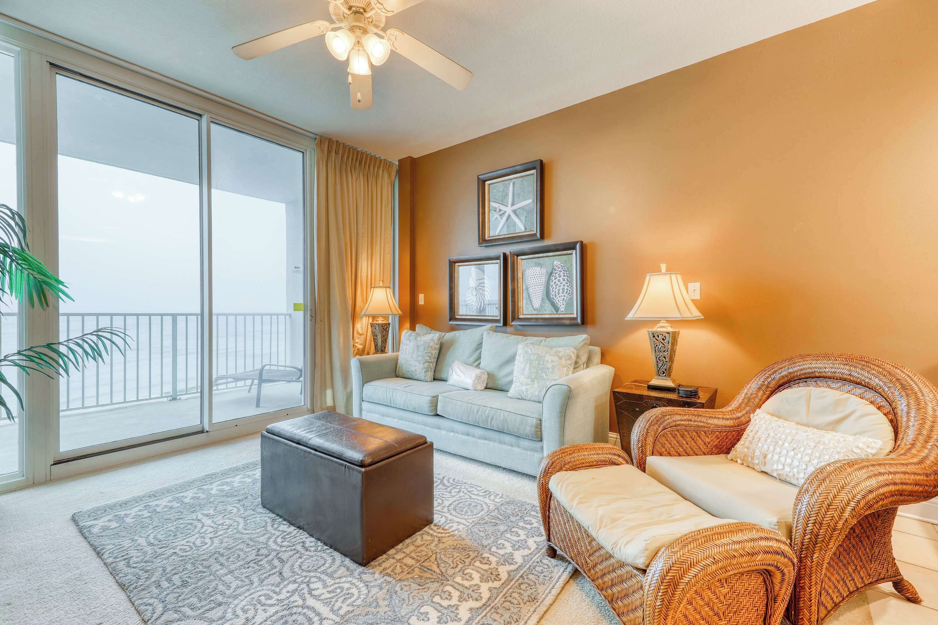 A living room with a sofa, ottoman, wicker chair, and a balcony with a view.