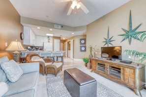 A living room with a sofa, a coffee table, a TV on a wooden cabinet, and a ceiling fan. - Beachfront condo w/ balcony, indoor & outdoor pools, sauna & hot tubs (Gulf Shores)