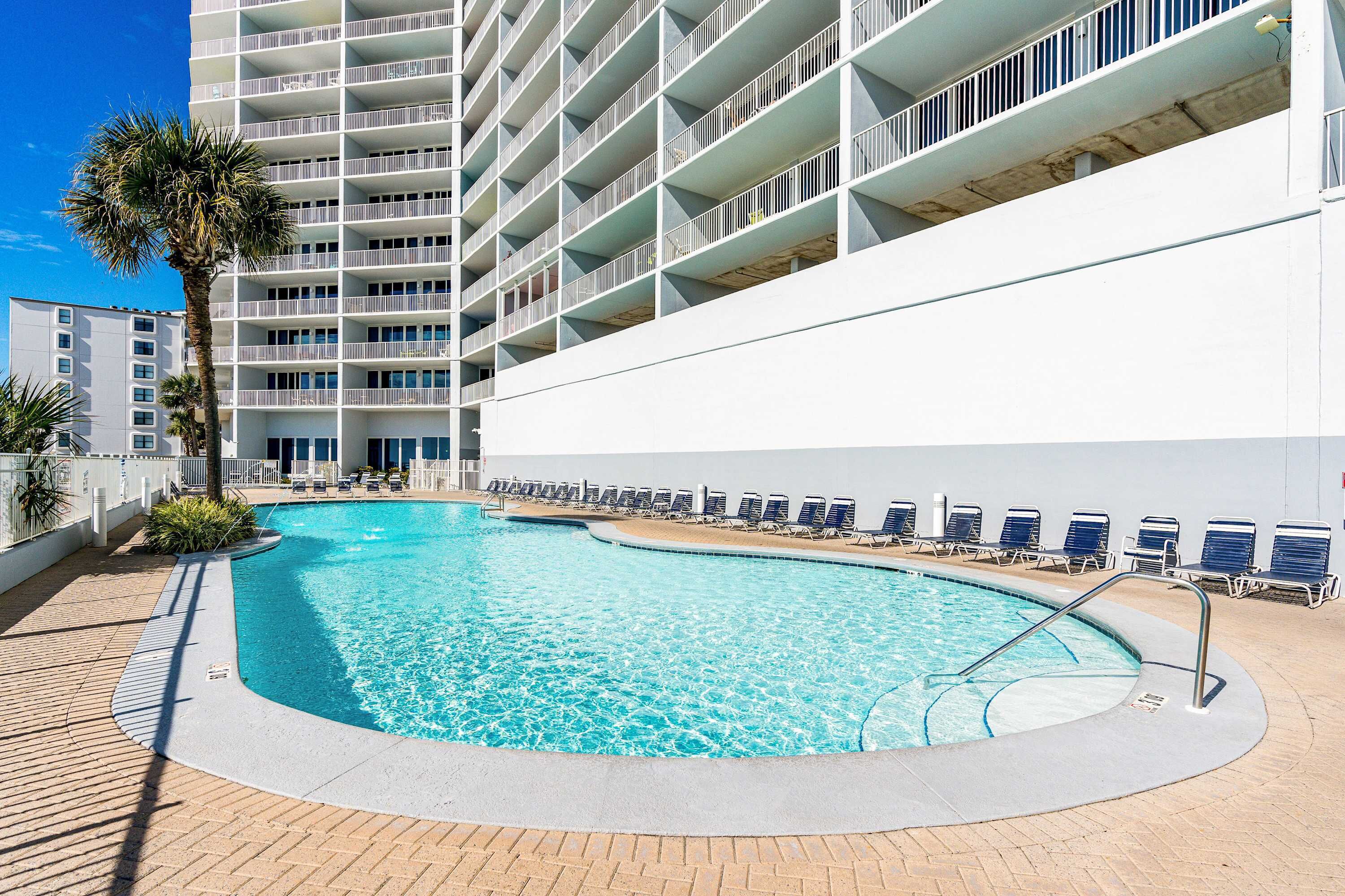 A swimming pool with lounge chairs, a palm tree, and a multi-story building with balconies.