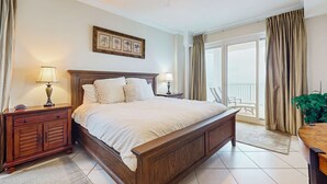 Ein Schlafzimmer mit einem großen Bett, Nachttischen aus Holz, einem Deckenventilator, einem Balkon mit Vorhängen und einem gerahmten Bild an der Wand.