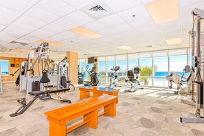 Un gimnasio con diversos equipos de ejercicio, un banco de madera y grandes ventanales con vista a la playa.