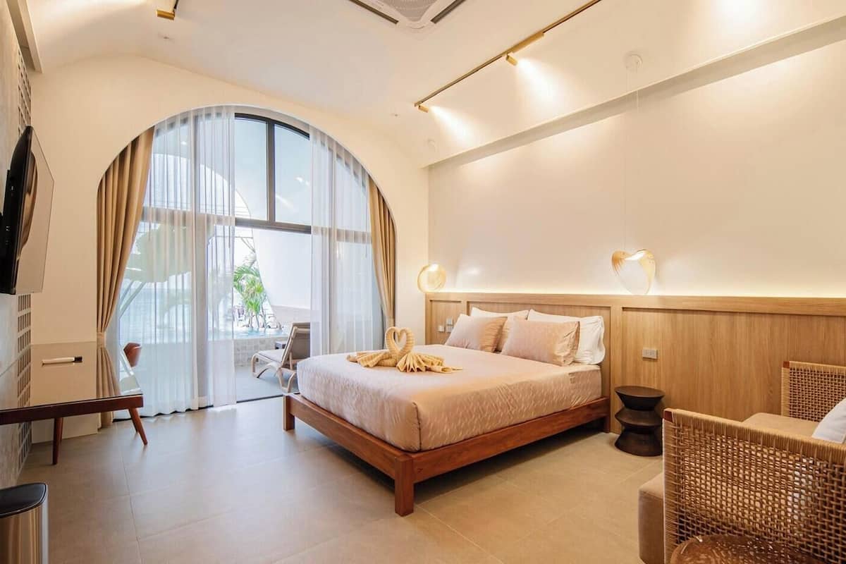 A modern hotel room with a large bed, a wicker chair, a desk, and a view of the outdoors.