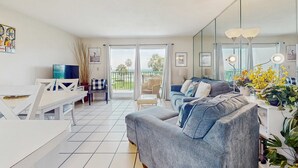 A modern living room with a grey sofa, a dining table, and a balcony with a view of greenery. - Waterfront condo with shared pool & hot tub - near beach at Plantation Palms (Fort Morgan)