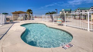 A swimming pool with a white fence, a palm tree, and a building with a green roof in the background. - Waterfront condo with shared pool & hot tub - near beach at Plantation Palms (Fort Morgan)