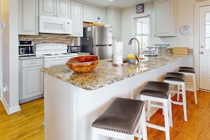 A kitchen with white cabinets, a granite countertop, a large island with stools, a stainless steel refrigerator, and a microwave. - Dreamy, dog-friendly, coastal retreat with screened porch - walk to the beach (Fort Morgan)