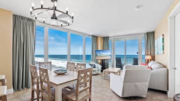 A room with a dining table, chairs, a sofa, a TV, and a view of the beach.