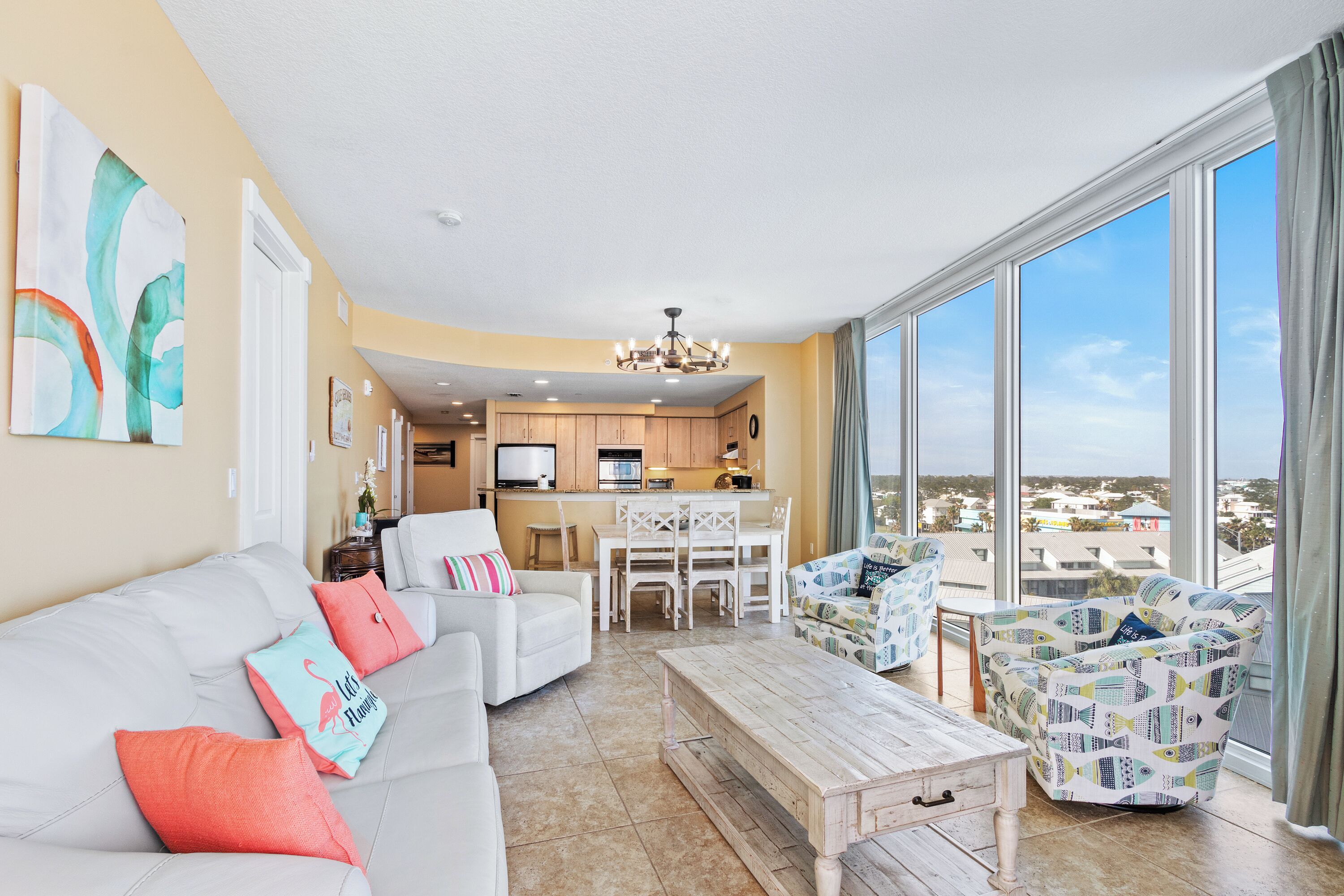 A spacious living area with a sofa, coffee table, and patterned armchairs, a kitchen in the background, and large windows offering a view.