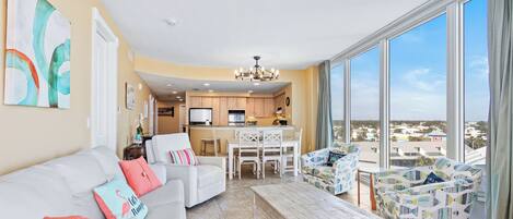 A spacious living area with a sofa, coffee table, and patterned armchairs, a kitchen in the background, and large windows offering a view.