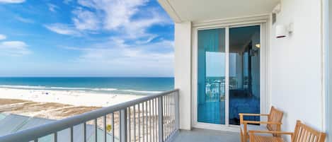 A balcony with a view of the beach, wooden chairs, and a glass door leading to an interior space.