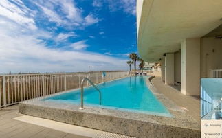 Piscina con vista a la playa y palmeras, rodeada por una valla.