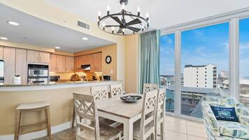 A modern kitchen with wooden cabinets, a dining area with a wooden table and chairs, a large window with a city view, and a ceiling fan.