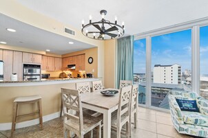 A modern kitchen with wooden cabinets, a dining area with a wooden table and chairs, a large window with a city view, and a ceiling fan.