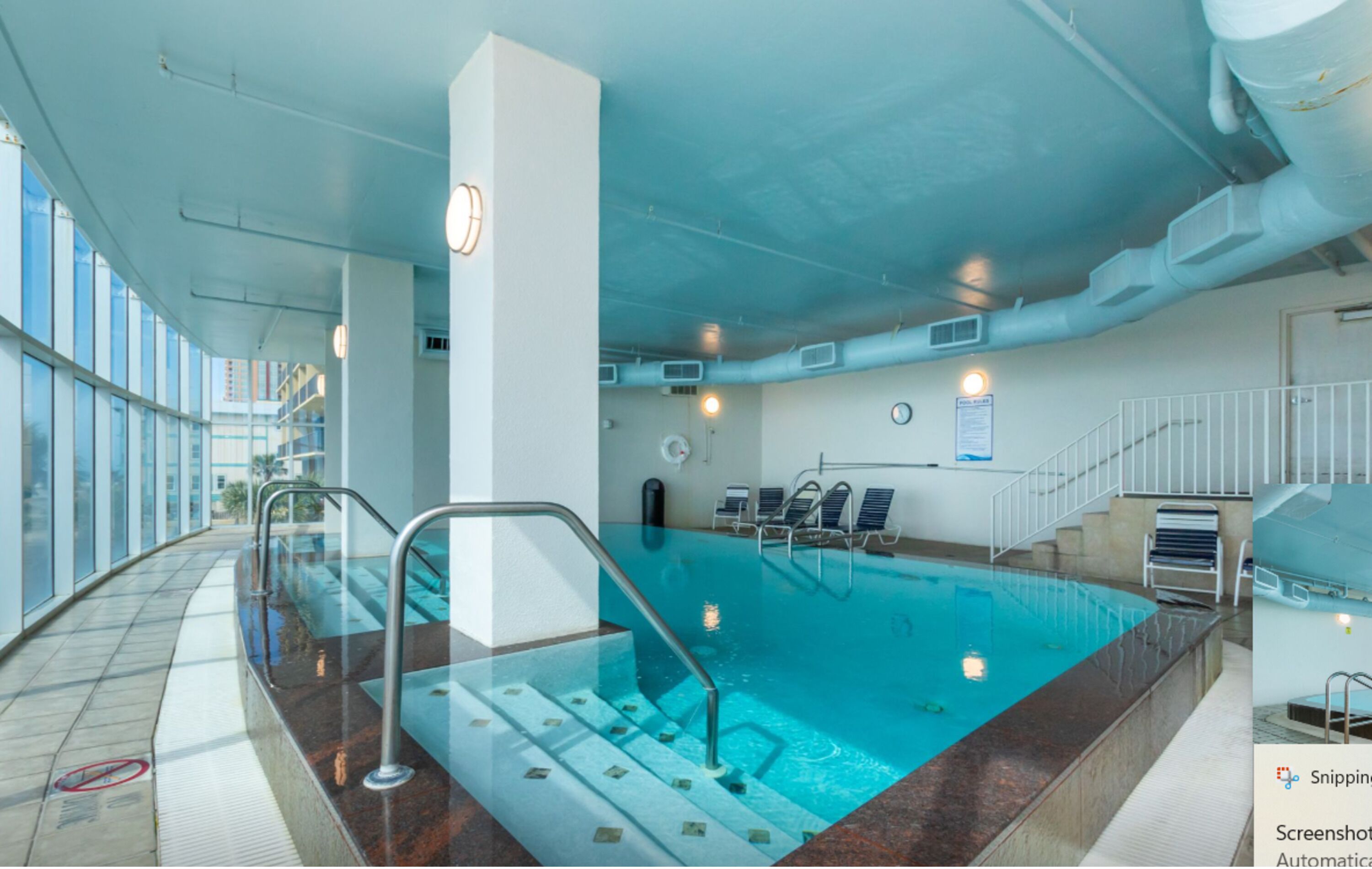 A modern indoor swimming pool with clear glass walls, a tiled floor, and a metal ladder leading into the pool.